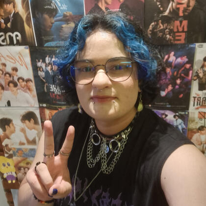 Selfie minha (Alex). Sou uma pessoa não-binária branca, tenho cabelo curto/médio, azul na frente e preto atrás e tenho 3 piercings no rosto. Estou usando uma regata preta, um choker preto com spikes e correntes e estou fazendo o símbolo da paz.